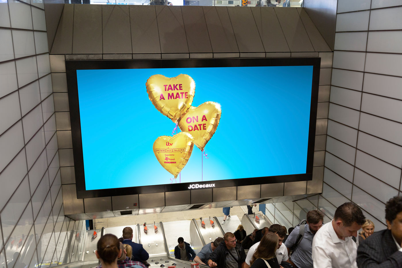 Displayed on a screen above a set of underground escalators is an image of three gold heart-shaped balloons on a blue background. The first two balloons have pink capital letters printed on them that read 'take a mate on a date'. The third balloon has an ITV logo, the Britain Get Talking logo and the text 'supported by Mind and YoungMinds'. 