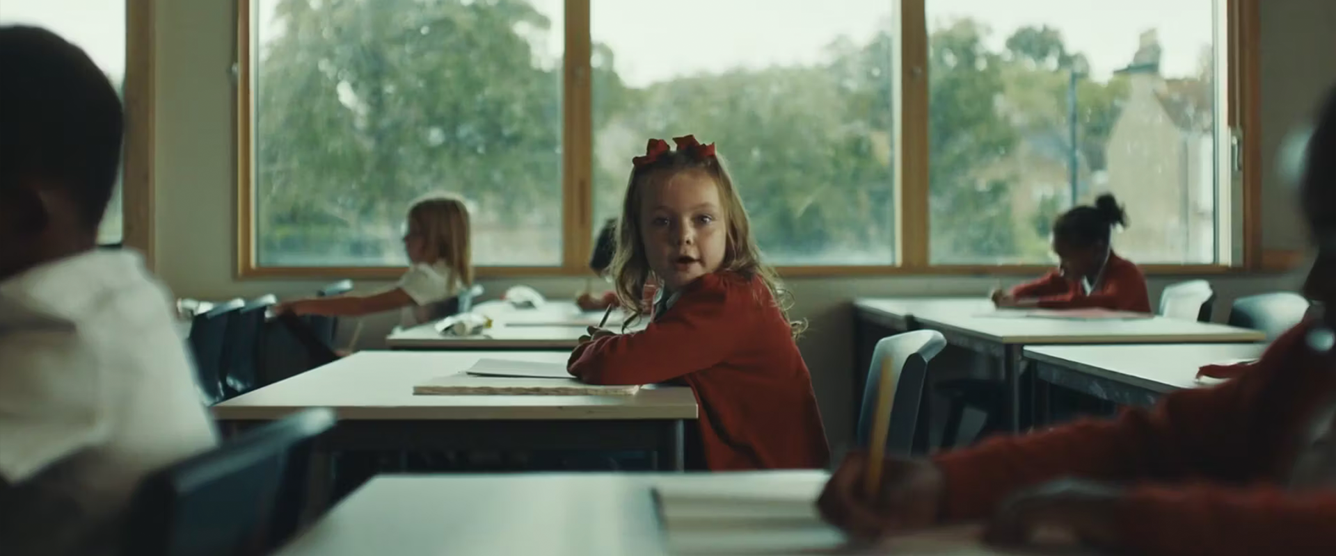 A young girl dressed in school uniform is sitting at a desk in the classroom surrounded by other pupils. She has two ribbons in her hair. She is looking away from the work on her desk towards the camera.