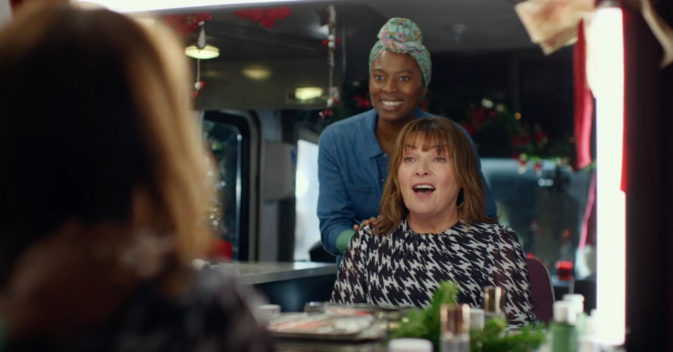 ITV presenter Lorraine is sitting in a make up chair in a TV production trailer. She is chatting. Behind her there is a make up artist listening and smiling.
