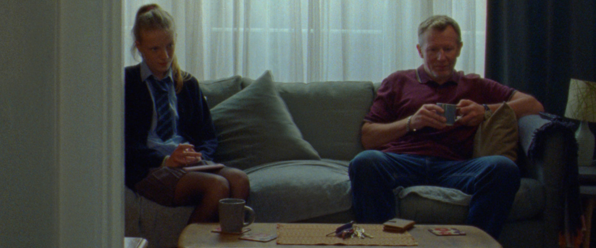 A young girl in a school uniform is sitting on a sofa next to her dad, who is holding a cup of tea. They are both looking down rather than communicating with one another.